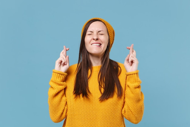 Young woman in sweater hat posing isolated on blue background studio portrait. People lifestyle concept. Mock up copy space. Wait for special moment, keeping fingers crossed, eyes closed, making wish.