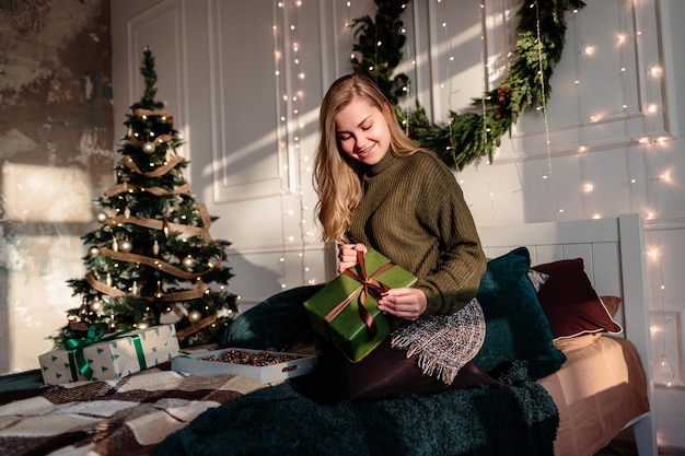 A young woman in a sweater opens Christmas gifts in a bedroom decorated for Christmas against the backdrop of a Christmas tree.