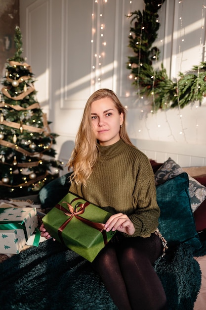 A young woman in a sweater opens Christmas gifts in a bedroom decorated for Christmas against the backdrop of a Christmas tree.