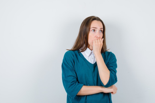 Young woman in sweater over white shirt biting nails while looking away and looking anxious , front view.