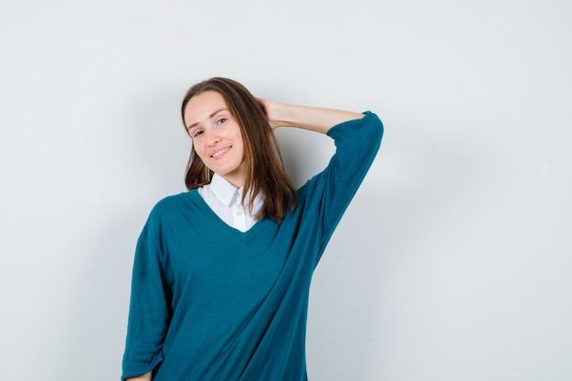 Young woman in sweater over white shirt posing with hand behind head and looking cheery , front view.