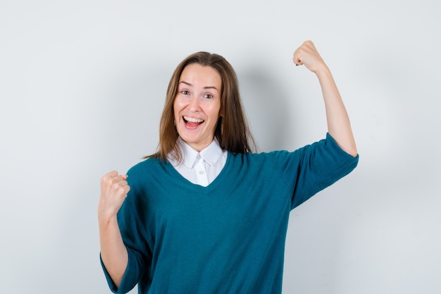 Young woman in sweater over white shirt showing winner gesture and looking happy , front view.