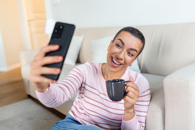 A young woman in a sweater with a cup in her hands looks at the phone while sitting on the couch