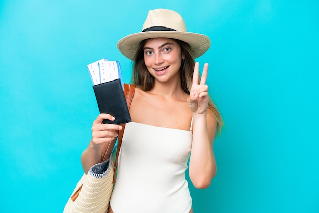 Young woman in swimsuit holding passport isolated on blue background smiling and showing victory sign