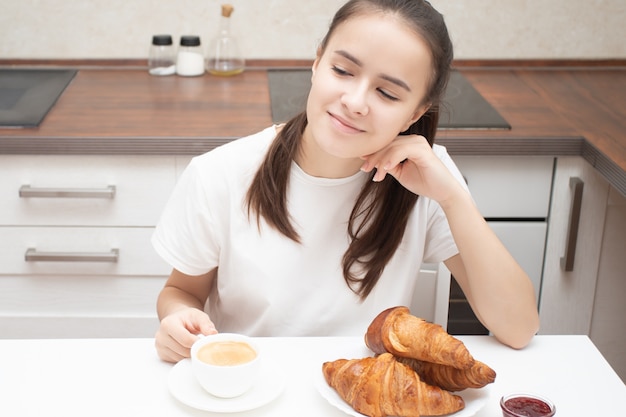 A young woman at a table in the kitchen having breakfast, smiling, talking, eating fresh croissants. Is drinking coffee