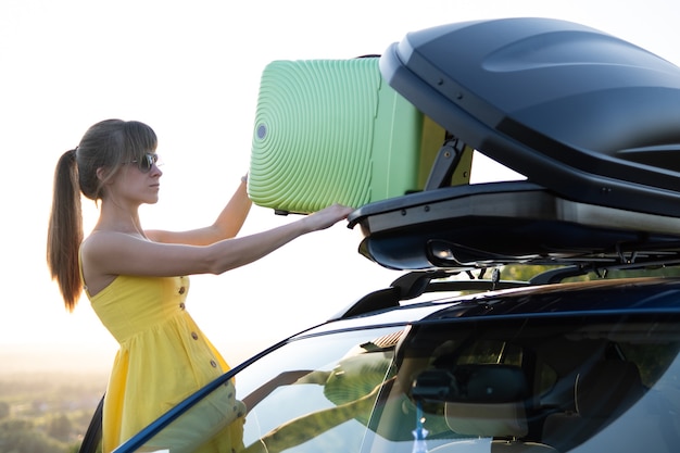 Young woman taking green suitcase from car roof rack. Travel and vacations concept.