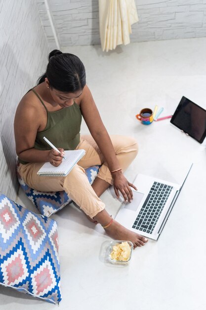 Young woman taking notes sitting on the floor with laptop and coffee near window