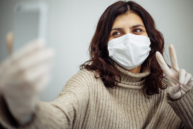 Young woman taking selfie photogragh and working from home during quarantine due to coronavirus pandemia. Beautiful girl stays home wearing medical mask and gloves. Covid-19 epidemia concept.