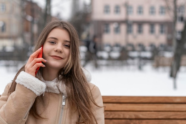 Young woman talks on phone sits on park bench Smiling young woman in beige sheepskin coat rests outside in winter