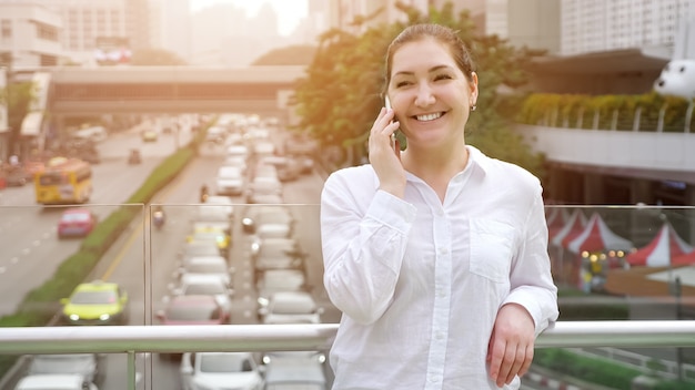 Young woman talks on white smartphone and smiles cheerfully standing on bridge over street between modern city buildings close up