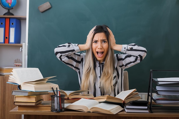 young woman teacher sitting at school desk in front of blackboard in classroom with books looking at mess on her desk being crazy mad shouting in panic holding hands on her head