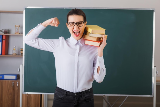 Young woman teacher wearing glasses explaining lesson holding stack of books crazy happy raising clenched fist standing at school desk in front of blackboard in classroom