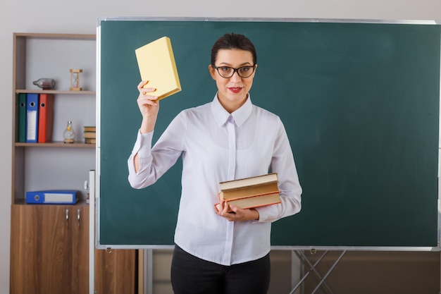 Young woman teacher wearing glasses explaining lesson holding stack of books smiling confident standing at school desk in front of blackboard in classroom