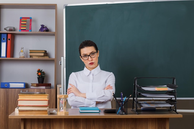 Young woman teacher wearing glasses explaining lesson looking at camera with serious face frowning crossing hands on chest sitting at school desk in front of blackboard in classroom