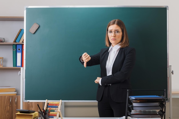 young woman teacher wearing glasses standing near blackboard in classroom explaining lesson showing thumb down being dissatisfied