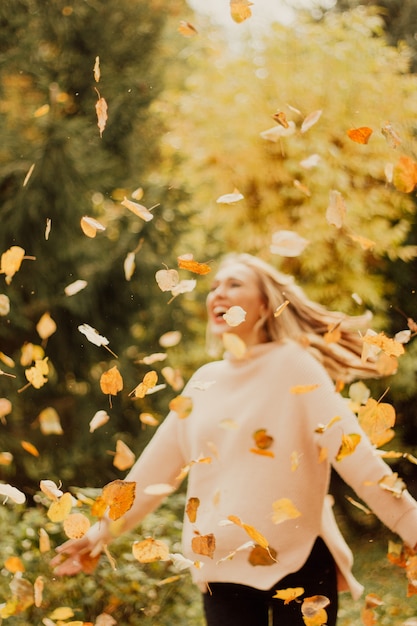 Young woman throws autumn yellow leaves. Happy girl in a sweater playing with yellow leaves outdoors.