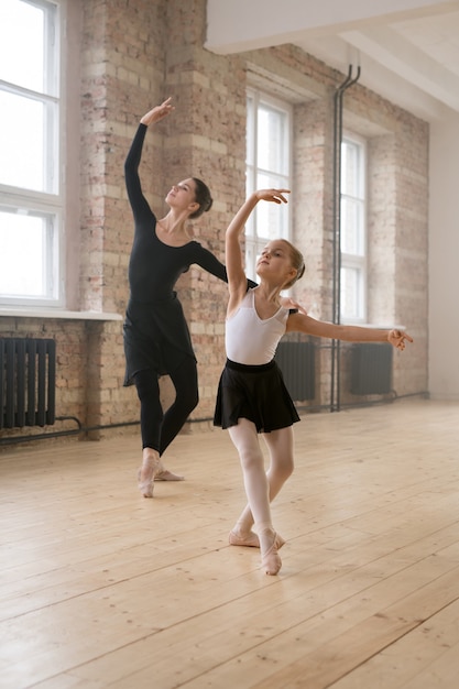 Young woman together with little girl dancing ballet during their training in dance studio