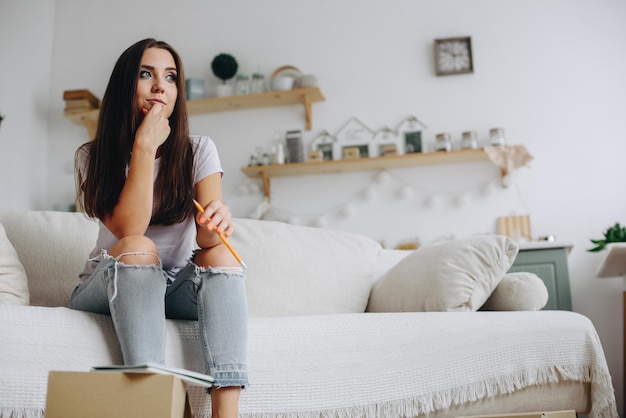 A young woman in torn jeans sits pensively on a sofa Pencil in hand Concept Unemployed