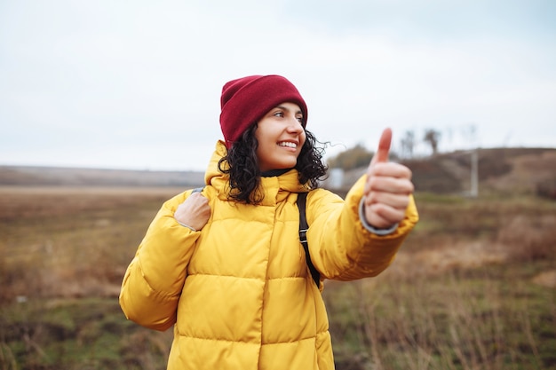 Young woman tourist catches a car on the winter road far from the city. Focus on face. Female wearing yellow jacket, backpack and a red hat.