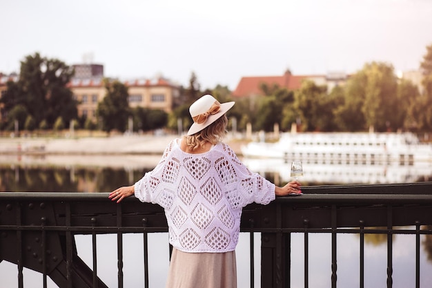 Young woman tourist standing with glass of white wine at bridge in european city river at background