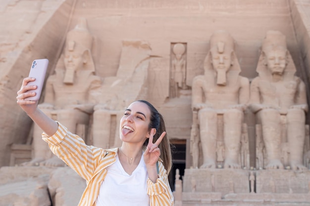 Young woman tourist taking a selfie with tounge out and peace out sign in Abu Simbel temple Egypt