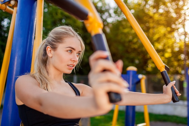 Young woman training on machine on sportsground in summer. Working on biceps muscles. Sportive lifestyle