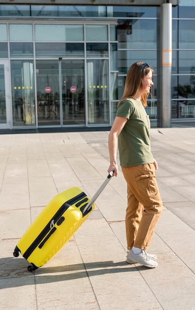 Young woman traveler in casual clothes carrying a yellow suitcase next to the entrance to the airport outside tourism concept of air flights travel and vacation