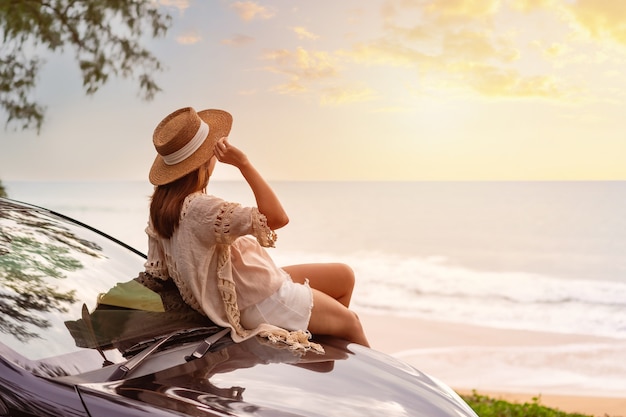 Young woman traveler sitting on a car and looking a beautiful sunset at the beach while travel driving road trip on vacation