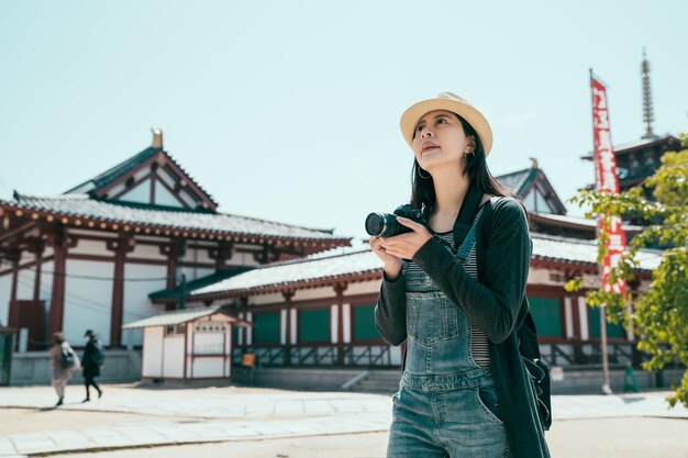 young woman traveler taking photo at temple in japan. asian lady tourist in denim overalls using camera take picture of shitennoji in osaka. female backpack photograhper standing under sunshine.