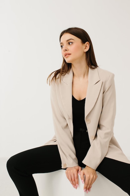 Young woman in trendy pastel suit sitting on cube against white background