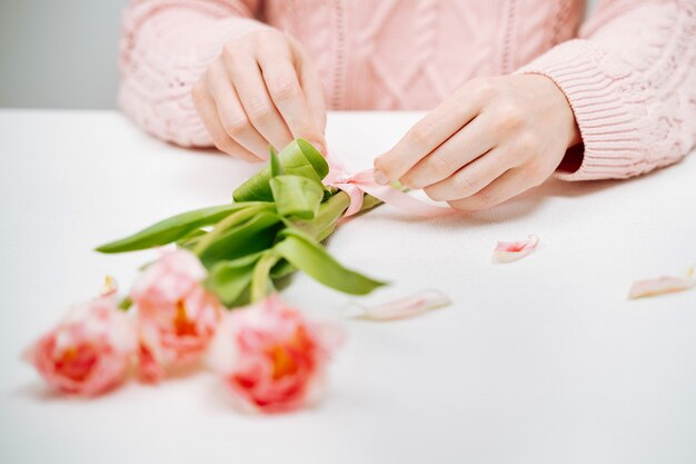 Young woman tying a ribbon on a bouquet of pink tulips. Selective focus, white background, text copy space.