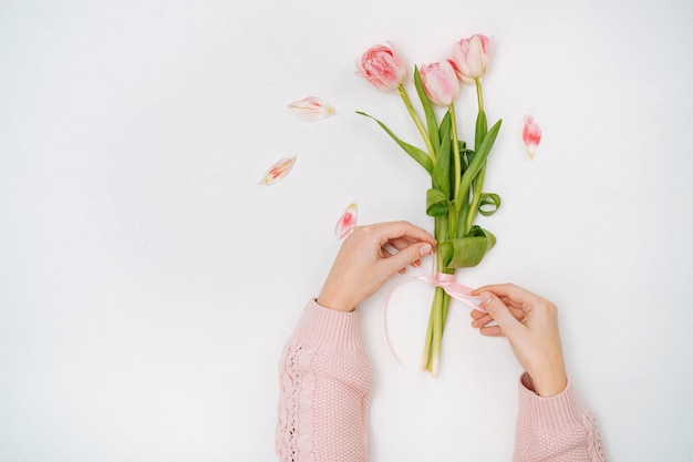 Young woman tying a ribbon on a bouquet of pink tulips. Top view, white background, text copy space. 8 March or Mother's Day.