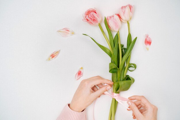 Young woman tying a ribbon on a bouquet of pink tulips. Top view, white background, text copy space.