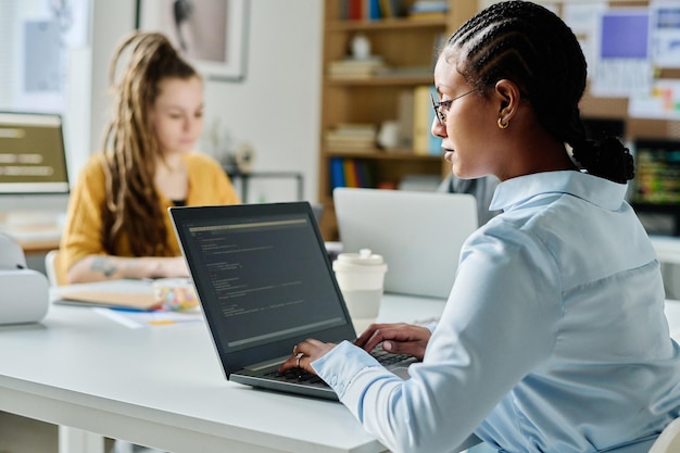 Young woman typing codes on laptop while sitting at table with her colleague in office