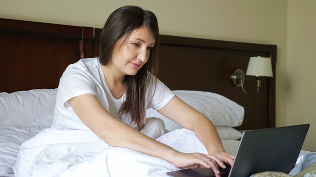 Young woman typing on a laptop while sitting in bed covered with a blanket