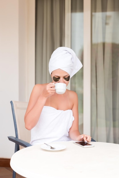 Young woman in undereye patches writing sms message and drinking coffee at hotel terrace resort morning holiday concept