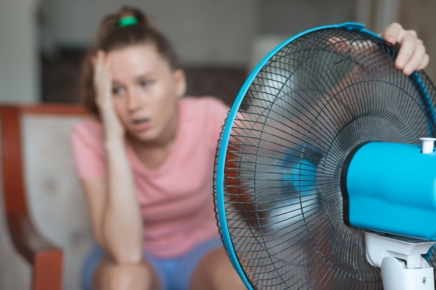 Young woman using electric fan at home in living room sitting on couch cooling off during hot