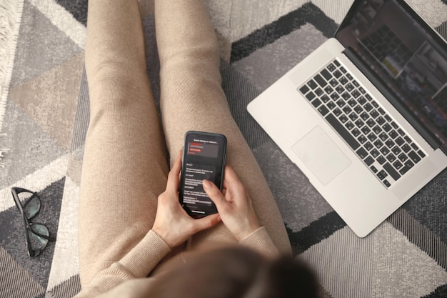 Young woman using her mobile phone while working with laptop sitting on the floor at home