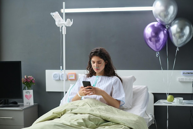 Young woman using her smartphone while lying on bed in hospital ward decorated with balloons