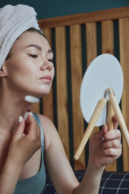 Young woman using jade facial roller for face and neck massage sitting on bed in bedroom looking in the mirror