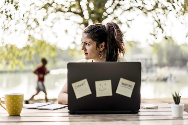 Young woman using laptop computer in the public park Millennial female freelancer working remotely and taking notes in a city park Technology and remote work concept Using tech devices outdoors