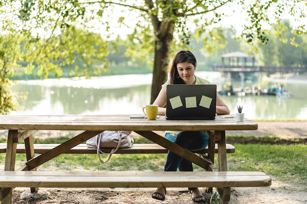 Young woman using laptop computer in the public park Millennial female freelancer working remotely and taking notes in a city park Technology and remote work concept Using tech devices outdoors