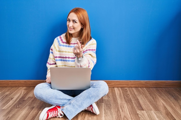 Young woman using laptop at home sitting on the floor beckoning come here gesture with hand inviting welcoming happy and smiling