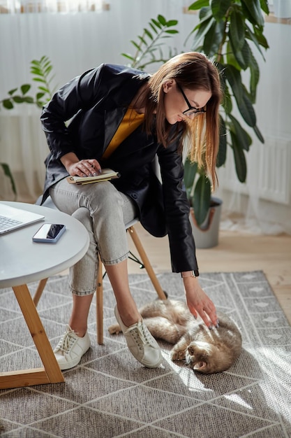 Young woman using laptop and mobile phone stroking a pet cat at workplace at home remote work