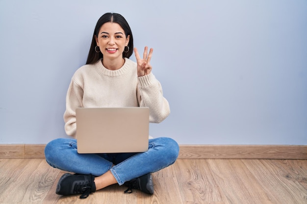 Young woman using laptop sitting on the floor at home showing and pointing up with fingers number three while smiling confident and happy