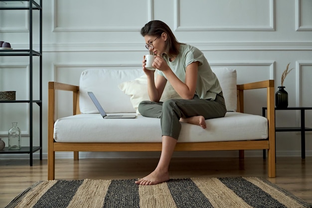 Young woman using laptop sitting at home on sofa and drinking coffee remote work work at home