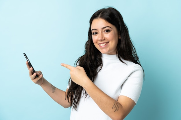 Young woman using mobile phone isolated on blue wall and pointing it