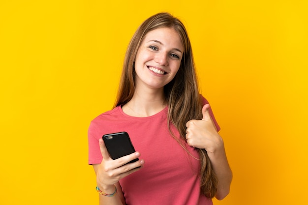 Young woman using mobile phone isolated on yellow with thumbs up because something good has happened