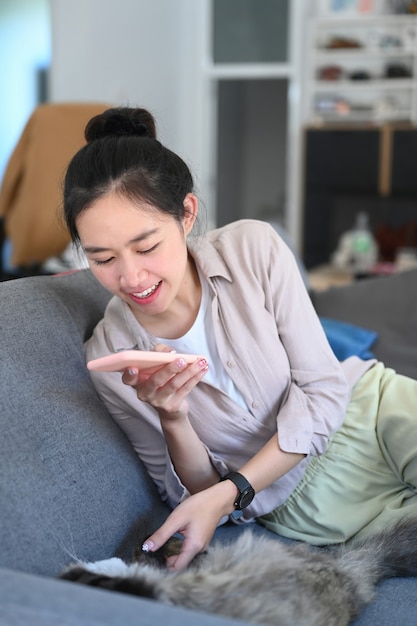 Young woman using smart phone taking photo of her cat on sofa in living room.