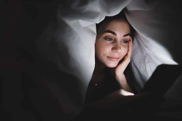 Young woman using a smartphone under a blanket on a bed